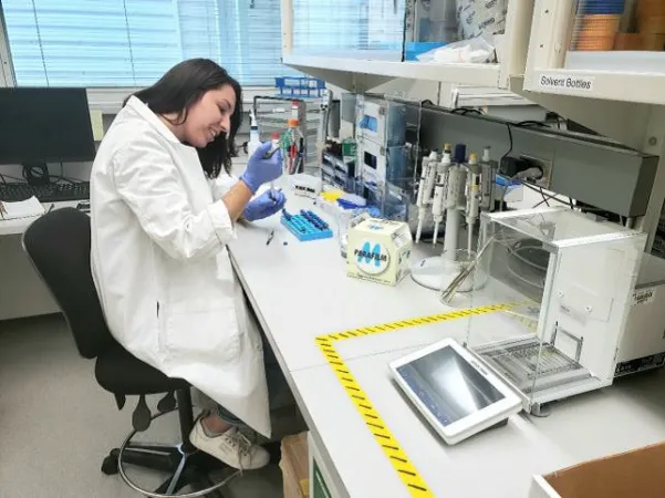 A young woman wearing a white lab coat and blue rubber gloves sits in a laboratory, working with a variety of tools in front of her. 
