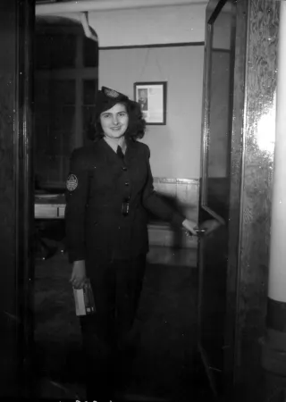 L’image est une photo noir et blanc montrant une femme en uniforme. Elle vient d’ouvrir une porte et s’apprête à passer le seuil, un télégramme à la main. 
