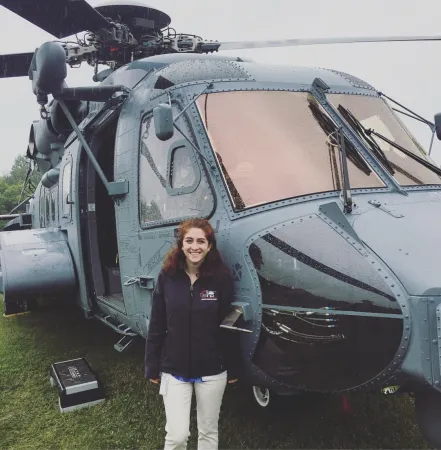 A young woman smiles as she stands next to a large helicopter, which is sitting on the grass.