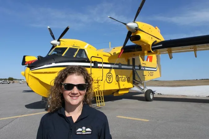 Une jeune femme portant des lunettes de soleil sourit à l’objectif de l’appareil photo. Derrière elle sur la piste se trouve un gros avion jaune, sur le côté duquel on distingue clairement le mot « Ontario ».