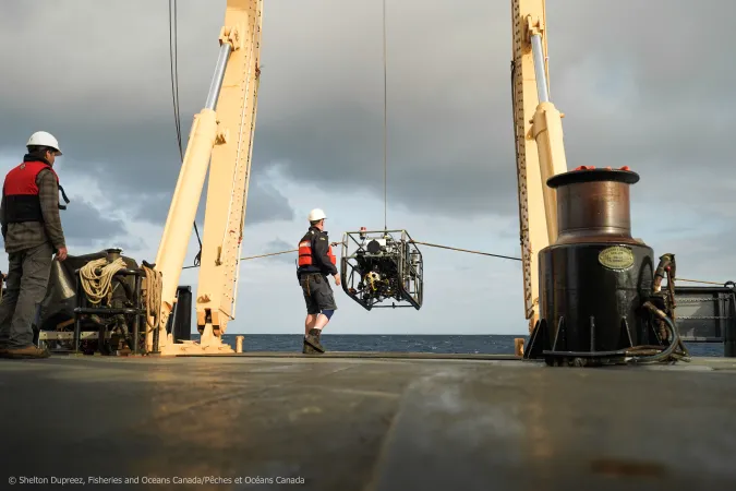 A crane on the back of a ship lowers an deep sea exploration robot while two workers observe