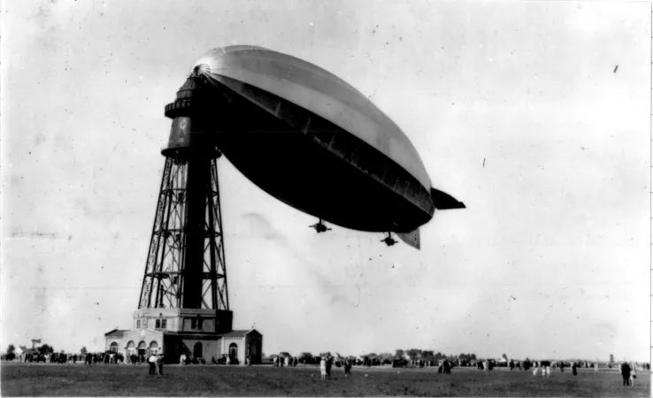 Une photographie noir et blanc montre de nombreuses personnes au-dessus desquelles un immense dirigeable, le R-100, est amarré au sommet d’une haute tour qui ressemble à un phare.