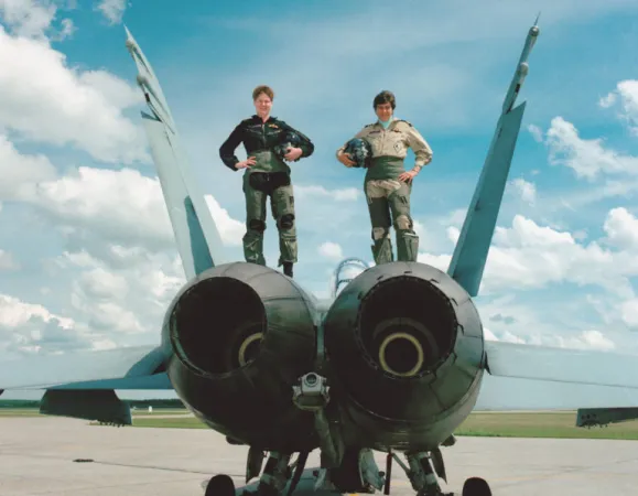 Deux femmes militaires en combinaison de vol posent avec confiance au sommet d'un avion de chasse sur une piste d'atterrissage par une journée claire et bleue. 