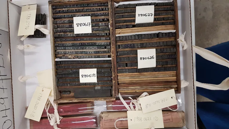 A white box is filled with seven objects. Two of them are boxes of metal type, labelled with white tags and black writing. The labels read: “880617,” “880618,” “880627,” and “880628.” The boxes are surrounded by the other artifacts that they will be packed with for transfer.  