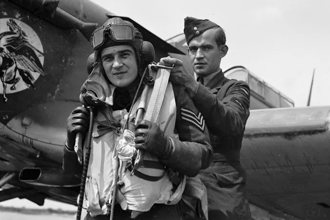 A black-and-white image depicts two men in military uniforms, standing in front of an aircraft. The man in front is a pilot, and is being helped into his parachute by the man standing behind him.