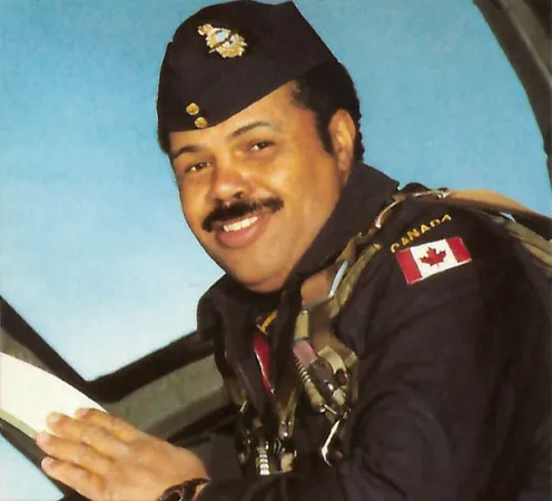 Une image en couleur montre un homme noir vêtu d’un uniforme d’aviateur posant à l’intérieur de la cabine de pilotage d’un avion. 
