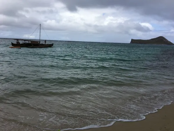 Image of an outrigger boat near the coast in open water in Hawaii with some sand visible on the shore and a small island in the distance.