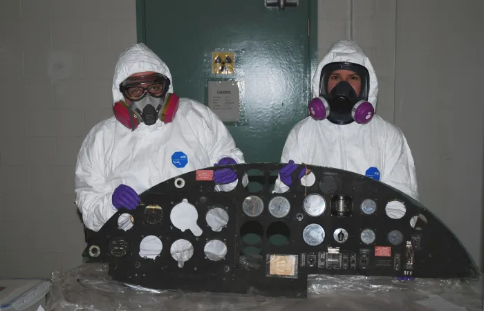 Two museum conservators wearing full-body Personal Protective Equipment stand side by side, holding a black aircraft control panel upright. The artifact is sitting on plastic sheeting on a table. 
