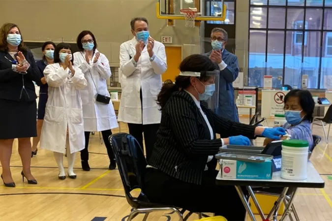 Une femme reçoit un vaccin contre la COVID-19 qui lui est administré par une travailleuse de la santé dans un gymnase d’école. Debout, à l’arrière-plan, six personnes regardent la scène et applaudissent.