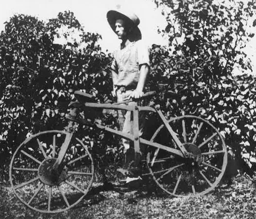 A black-and-white photo of a man standing outside with his left hand on the seat of a wooden bicycle.