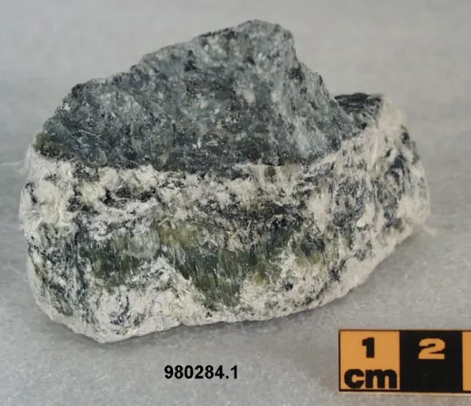 L’amiante à l’état de minerai. Le minéral est verdâtre et blanc, et on aperçoit des fibres qui s’en détachent.  