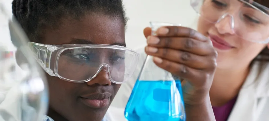 Un gros plan sur deux jeunes personnes portant des lunettes de protection dans un milieu évoquant un laboratoire. La personne au premier plan tient un bécher en verre rempli d’un liquide bleu.