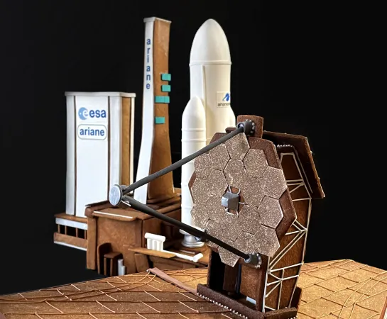 Two gingerbread and sugar creations are pictured against a dark backdrop; a model of the James Webb Space Telescope is in the foreground, and the Ariane 5 rocket is visible in the background.