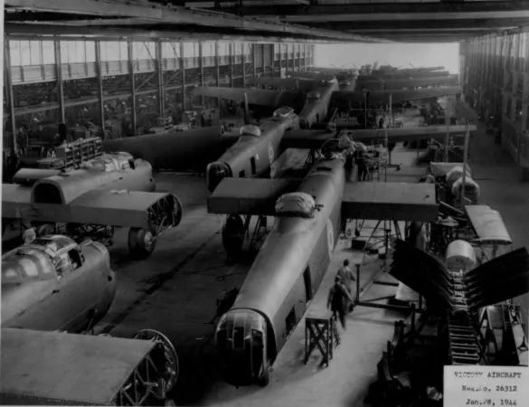 Photo en noir et blanc d’une dizaine d’avions en construction, dans une grande usine. Deux ouvriers sont visibles en avant-plan.