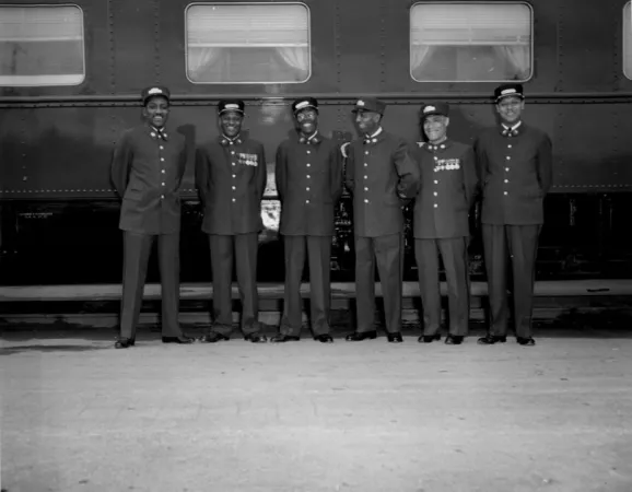 Photo noir et blanc de six porteurs noirs en uniforme, côte à côte devant un train. Souriants, ils regardent la caméra ou se regardent entre eux.