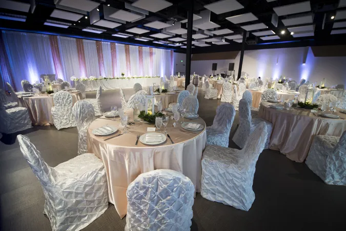 Une grande pièce avec des rideaux blancs et roses et plusieurs tables rondes avec des nappes roses qui tombent jusqu’au sol et des chaises somptueusement drapées de blanc. Les tables sont dressées de façon élégante et les luminaires projettent un éclairage bleuté.
