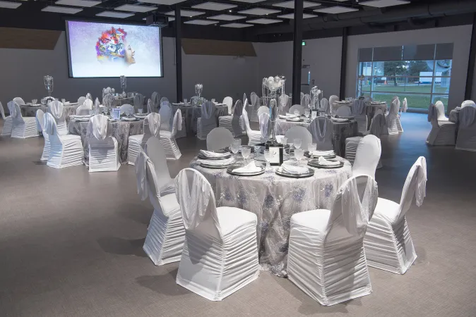 Une grande pièce aux murs blancs, avec plusieurs tables rondes recouvertes de nappes blanches qui tombent jusqu’au sol et des chaises somptueusement drapées de blanc. Les tables sont dressées de façon élégante, avec une assiette et des ustensiles argentés à chaque place. Un grand écran est installé sur un des murs.