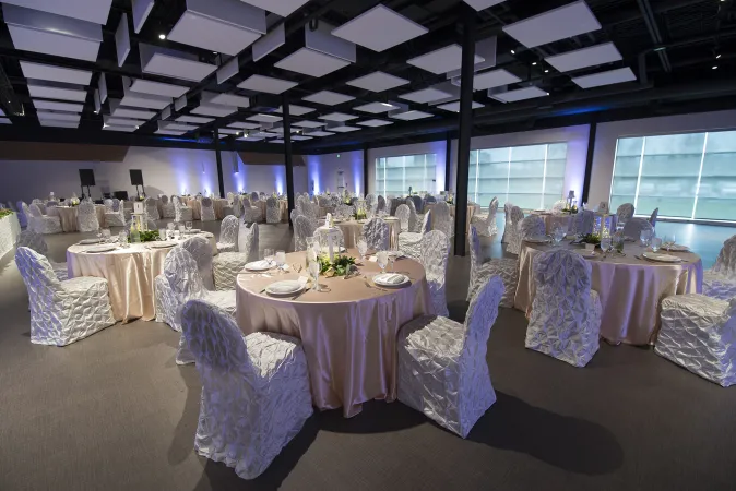 Une grande pièce avec des rideaux blancs et roses et plusieurs tables rondes avec des nappes roses qui tombent jusqu’au sol et des chaises somptueusement drapées de blanc. Les tables sont dressées de façon élégante et les luminaires projettent un éclairage bleuté.