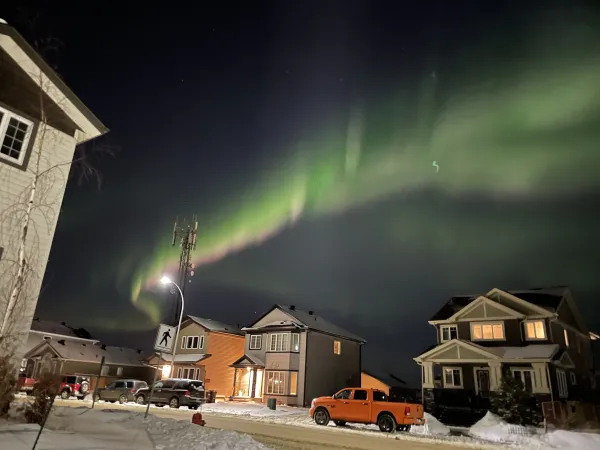 Green and pink Northern Lights glow above a snowy residential street. An orange pickup truck is parked in front of one of the houses. 