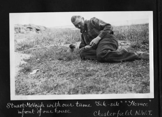Black and white horizontal photograph of man wearing glasses who is laying on his side on the ground feeding a small squirrel by hand. 