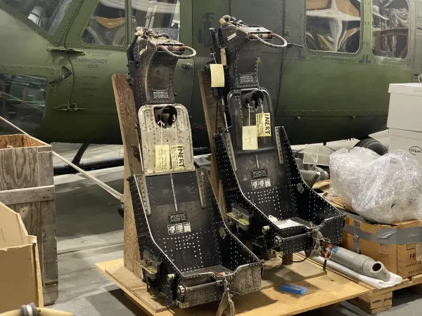 A pair of bare metal ejection seats with no cushions or padding are mounted on a plywood base, placed beside a few cardboard boxes. Behind the seats, a camouflage-green helicopter is partially visible.