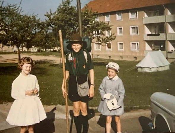 Three smiling children stand on a green lawn. On the far left is a young girl with short brown hair who wears a light pink dress. In the middle, a taller boy wears a scouting uniform. On the right, a young child wears a formal blue coat as well as a white hat, gloves, and purse.