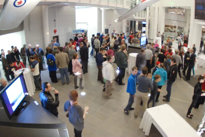 A view inside the Canada Aviation and Space Museum's lobby during an event. People are standing and networking. Cocktails tables are set up around the room with tablecloths.