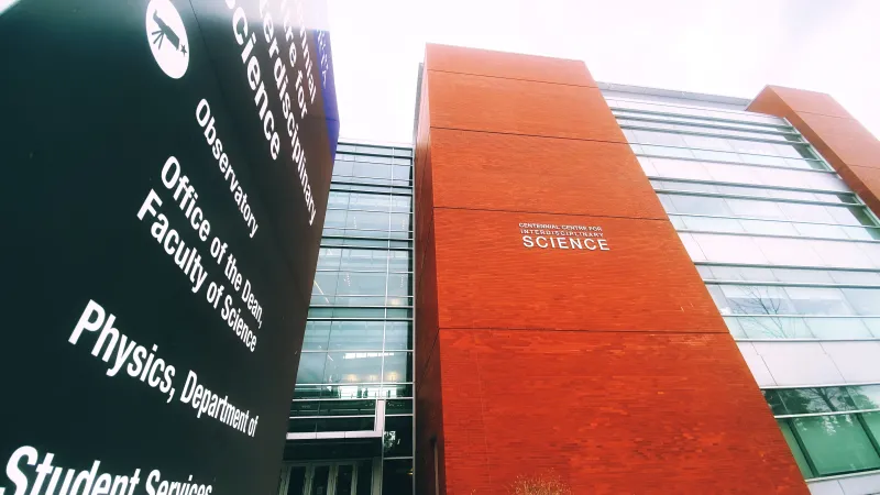 Low-angle view of signs on the University of Alberta campus. Left is a black wayfinding sign that reads “Physics, Department of” in white writing. Right is a white building sign that says “Centennial Centre for Interdisciplinary Science.” This sign is affixed to a multi-floor red brick exterior wall, with windows on either side.