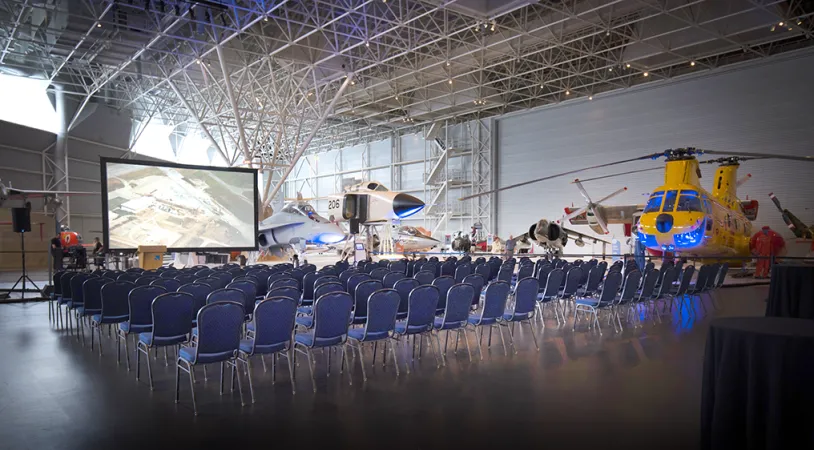 Hundreds of chairs lined up facing a projector and screen in the center of a large room. A yellow helicopter can be seen in the background along with part of a small white airplane.