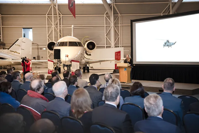 A large group of people are sitting in lines of chairs set up in the middle of a large room looking at a helicopter on a projected screen and listening to a person at the podium talking. In the background, multiple airplanes can be seen. 