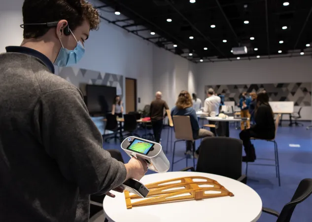 Une personne scanne en 3D la structure d'une aile d'avion dans une grande salle avec des tables et des chaises de différentes tailles. En arrière-plan, des personnes travaillent, certaines en groupe, d'autres individuellement.