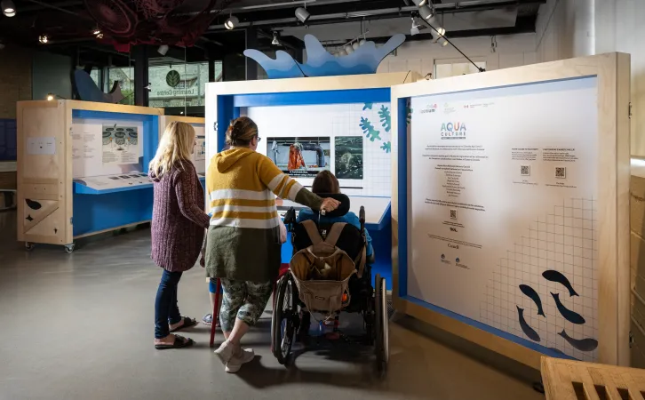 Three visitors, one of whom is using a wheelchair, interact with an exhibition video. On the screen, the inside of a salmon farm is shown.