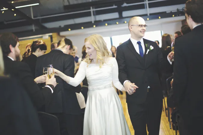 a bride and groom walking down the center aisle holding hands and smiling at the standing guests dressed in formalwear. 