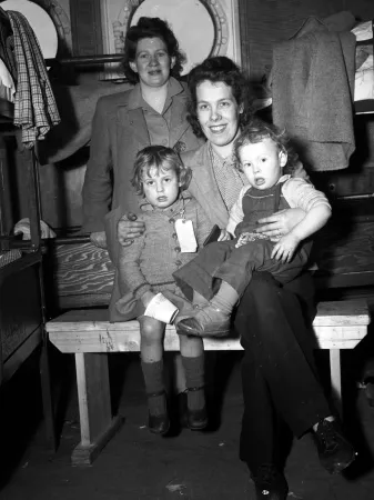 A black and white photograph of two women, one standing, one sitting on a bench, two small children sit with the woman on the bench. One child is wearing a tag on her coat.