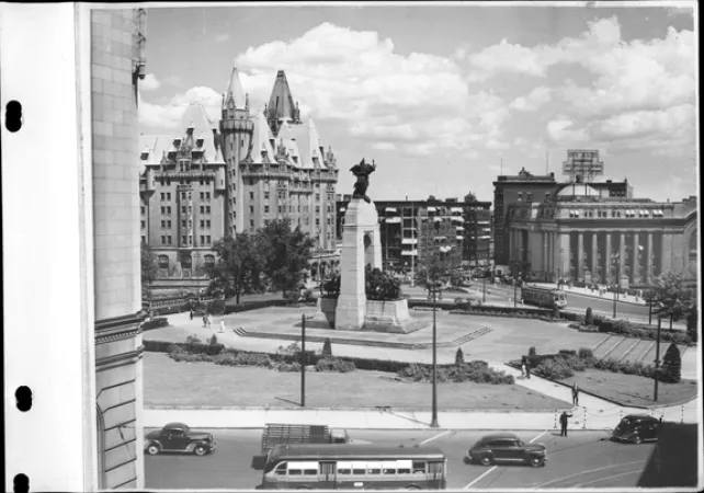 Image is a black-and-white photograph showing the War Memorial, the Château Laurier Hotel, and Union Station in Ottawa. The photograph is taken from a high perspective and shows buses and cars as well.  