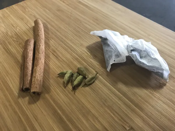 Two tea bags, two cinnamon sticks and a small pile of cardamom pods rest on a wooden butcher block.