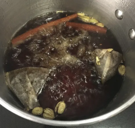 Une casserole de couleur argent est posée sur une cuisinière. À l’intérieur, on voit le thé infuser dans un liquide qui bouillonne. On voit des bâtons de cannelle, des gousses de cardamome et des sachets de thé dans l’eau bouillante.