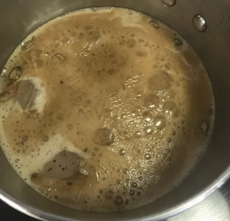 Une casserole de couleur argent est posée sur une cuisinière. À l’intérieur, le thé infuse dans un liquide laiteux qui mijote. À la surface, on voit flotter des sachets de thé parmi les bulles.