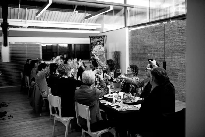 Black and white photo of a long table filled with people in formal dresswear. They are raising their glasses together.