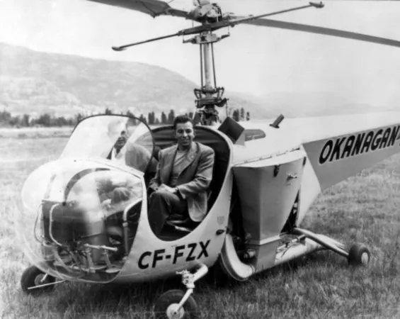 Image is a black-and-white photograph showing two men sitting in the open cockpit of the Bell 47B-3 helicopter. The helicopter is on the ground in a grassy field, and the word “Okanagan” is visible on its tail. 