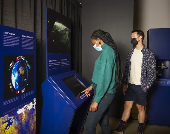 A person wearing a green top and black pants interacts with a touch screen on a blue museum module. To their left is a panel with an image of Earth. To their right a person looks on.