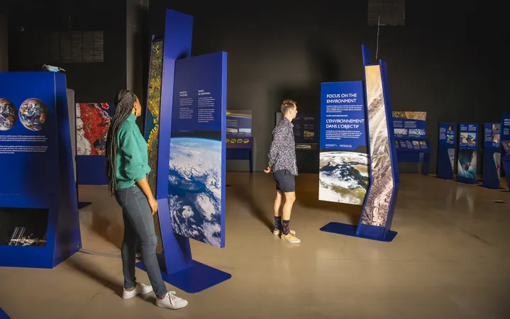 A wide view of an exhibition with two people reading text panels that include images of Earth taken from space.