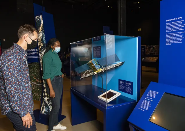 Two people are looking at a model of a satellite which is displayed in a large plexi glass display case with a blue base. In the foreground is a blue exhibition module with a touch screen.