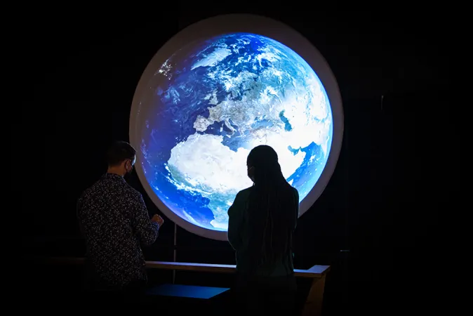 Two people stand in front of a large circular screen with an image of the Earth projected onto it.  The space around the screen is dark and the two people are in silhouette.