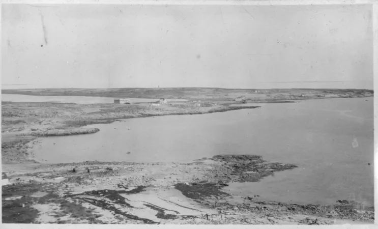 A horizontal black and white photograph of a harbor. The land is partially covered in snow and the sky is flat and clear. 