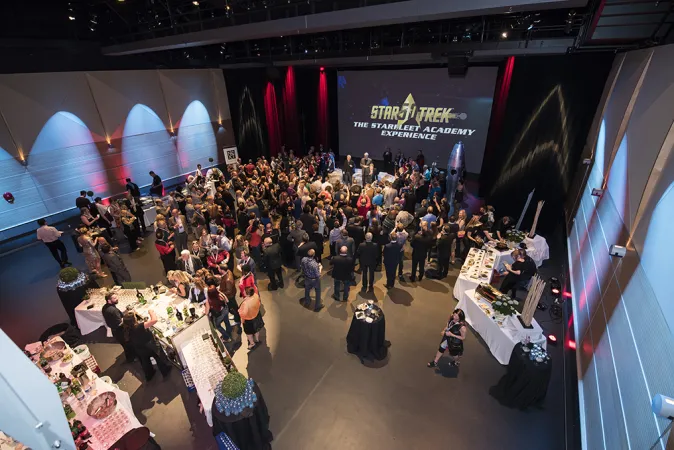 Top down view of a room decorated with various space-themed items and small circular black tables scattered around the room. Multiple tables are set up on one side of the room with food and drinks on the other side. On the projector screen, the words “Star Trek the startfleet academy experience” is displayed. Many people in formalwear are standing in groups around the room.