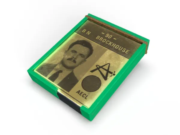 A plastic square with a green border. Inside the square is the black and white photo of a mustachioed man with suit and tie. Above the photo, in capital letters, is written: “- 90 – B N Brockhouse”, and what appear to be two logos one a circle, one resembling the capital letter A but drawn as if it’s a trajectory path of an aircraft, and the letters AECL.
