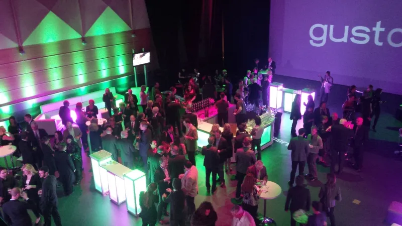 Many people talking in small groups inside of a large room with futuristic set up and décor. There is a screen projecting the word “Gusto” in purple on the wall and several small lit cubed tables and a drinks station. The walls of the room are lit up with green and purple lights.