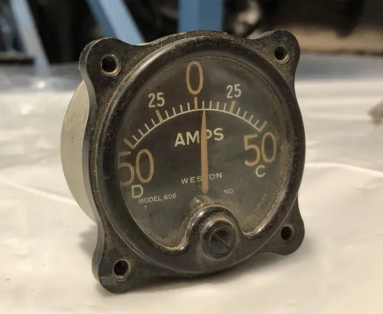Ammeter in a black-painted housing with a glass cover. The instrument face is black with white numbering and lettering. The scale reads from 50D through 0 to 50C “AMPS”. 