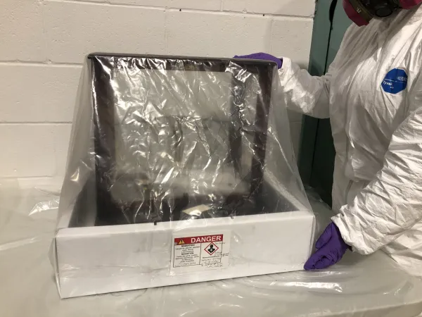 The balance is in a deep white tray and sealed in plastic. The tray has a hazard label tag marked “DANGER Radioactive”. An Ingenium conservator, wearing a Tyvek suit, respirator, eye protection and gloves, is holding the side of the balance.